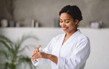 Beyaz cüppeli Afro-Amerikalı kadın, yeşil alanla dolu, parlak bir yerde ellerine losyon sürmekten mutluluk duyuyor. Güneş ışığı geniş pencerelerden geçer, sakin atmosferi güçlendirir..