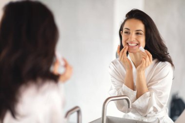 Sıcak bir banyo ortamında, esmer bir kadın yansımasına gülümserken cilt bakım ürünleri uyguluyor. Şımartıcı tavırları özgüven ve güzelliği vurguluyor..
