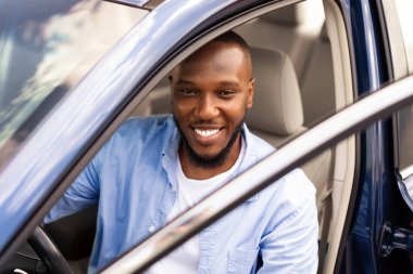 Sürücü koltuğunda oturan Afro-Amerikan bir genç. Açık otomobil kapısından kameraya bakıp gülümseyen mutlu siyah erkek. Otomatik test sürüşü, kiralama, satın alma, kiralama konsepti