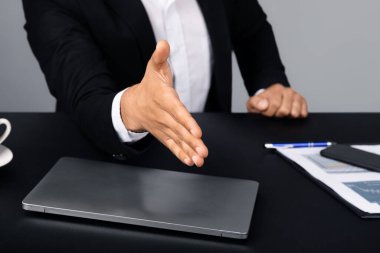 Cropped of entrepreneur in a suit offers a handshake across a desk with a laptop and documents. The setting reflects a modern corporate environment, emphasizing professional interaction. clipart