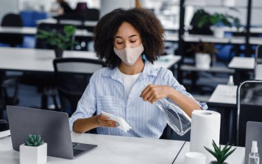 Koronavirüs ve yeni normallere karşı sağlığını korumak için işyeri hijyeni. Maskeli Afrikalı Amerikalı kadın ofis içinde masa ve laptopu dezenfekte etmek için sprey ve kağıt havlu kullanıyor.