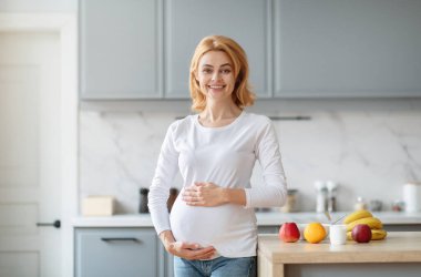 Hamile bir kadın modern bir mutfakta durur, bebeğinin karnını şişirirken ışıl ışıl gülümser. Taze elmalar, muzlar ve diğer meyveler tezgahın üzerinde sergilenerek neşeli bir atmosfer yaratılıyor..