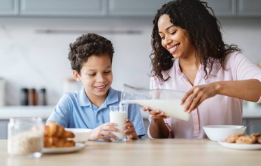 Sıcak bir mutfakta, bir anne sevgiyle oğluna süt döker, o da sevinçle gülümser. Birlikte özel bir an paylaşıyorlar, kahvaltı malzemeleri ve sıcaklıkla çevrili..