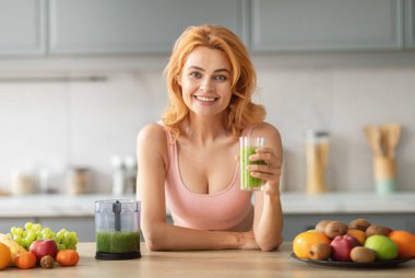 Neşeli genç bir kadın elinde yeşil bir meyve püresi tutarken gülümsüyor. Taze meyveler şık bir mutfakta, sağlıklı bir yaşam tarzı ve beslenme zevkini ön plana çıkarıyor..