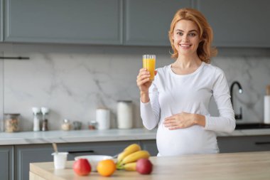 Hamile bir kadın şık bir mutfakta elinde neşeyle bir bardak portakal suyu tutuyor. Taze meyveler tezgahta sergileniyor ve evinde sağlıklı bir atmosfer yaratıyor..
