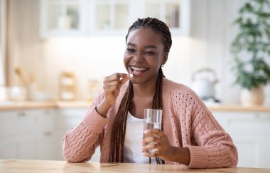 Güzellik malzemeleri. Vitamin hapı alan, evde su içen, gülümseyen, güzel cilt, saç ve tırnaklar için multivitamin tableti kullanan, kameraya bakan,