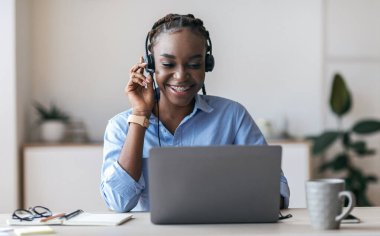 Neşeli siyah kadın yönetici kulaklık takıyor ve ofiste dizüstü bilgisayar kullanıyor, müşteriye danışıyor, müşteriyle konuşuyor, boş alan yaratıyor.