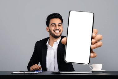 Genç bir Arap girişimci, modern bir stüdyoda masasında oturuyor, boş bir akıllı telefon ekranını tutarken güvenle gülümsüyor. O, resmi siyah bir takım elbise giymiş, başarılı bir yaşam tarzı yayıyor..