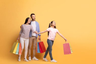 Üç kişilik mutlu bir aile, aralarında bir anne, baba ve genç kızın da bulunduğu, alışveriş yaparken gülümsüyor. Kız renkli çantalarla, gösterişli aile bağları ve sevgisi ile önde gidiyor.