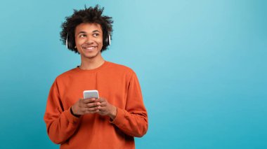 Kıvırcık saçlı genç bir Afro-Amerikan adam akıllı telefon tutarken müzik dinlemekten hoşlanır. Kulaklık takıyor ve parlak mavi bir arka plana karşı duruyor, neşe ve rahatlama ifade ediyor, boşluğu kopyalıyor.