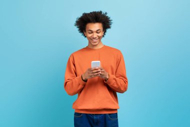 Gülümseyen Afro-Amerikan bir adam mavi bir arka plana karşı duruyor, akıllı telefonuyla meşgul. Turuncu bir kazak giyiyor ve rahat bir iç mekanda diğerleriyle bağ kurmaktan hoşlanıyor..