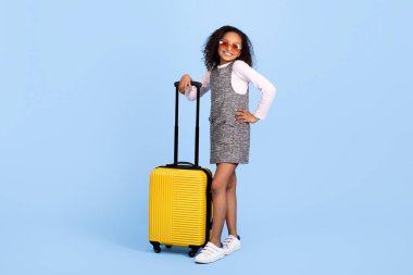 Neşeli siyah bir kız parlak sarı bir bavulla yaklaşan bir gezi için heyecanla bekliyor. Şık giysisi ve güneş gözlüğü, neşeli gençlik ruhunu canlı bir şekilde yansıtıyor..