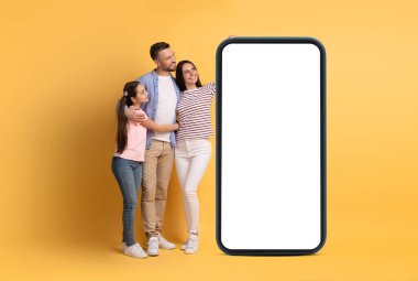 Üç kişilik bir aile, parlak sarı bir zemin önünde bir arada durur, baba ve genç kız annesine sarılırlar. Büyük bir akıllı telefonun yanında poz verirken gülümserler..