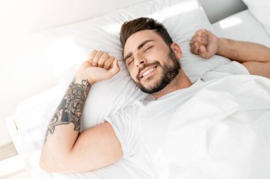 Mutlu uyuyan adam sabah uyandığında yatak odasında rahat bir yatakta uzanırken gülümsüyor ve pencereye bakıyor. Günaydın, uyanış konsepti..