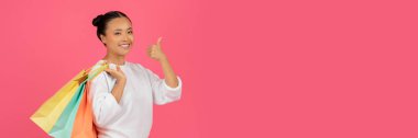 Büyük Alışveriş Konsepti. Renkli alışveriş torbaları tutan mutlu Asyalı kadının portresi ve başparmak hareketi gösteren neşeli bir kadın kameraya bakıyor, pembe arka planda tek başına duruyor,