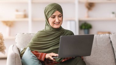 Neşeli, olgun, Müslüman bir kadın dizüstü bilgisayar kullanıyor. İnternette çalışıyor. Evde koltukta oturuyor. Modern Arap Serbest Çalışan Kadın Oturma Odasında İnternet Bilgisayarına Bakıyor. Serbest Kariyer Konsepti