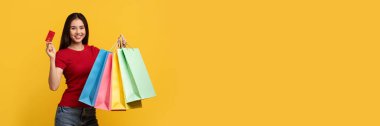 Renkli kağıt torbaları ve kırmızı plastik kredi kartı gösteren, kameraya gülümseyen, sarı arka planda izole edilmiş, mutlu, genç, çekici Çinli kadın müşteri. Kolay alışveriş, perakende, kopyalama alanı