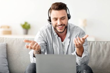 Çevrimiçi iletişim. Kulaklıklı mutlu bir serbest yazar dizüstü bilgisayarla video görüşmesi yapıyor, evde çalışıyor, kanepede oturuyor. Gülümseyen adam bilgisayarda web konferansı yapıyor, boş alan