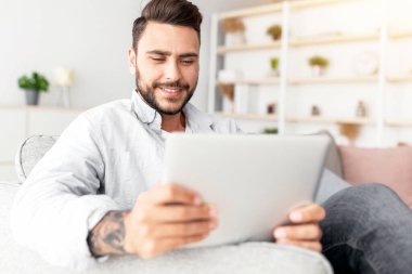 Beyaz bir adamın dijital tabletle kanepede dinlenmesine yakın çekim, ev içi, boş alan. Oturma odasındaki koltukta oturan mutlu adam, modern bloknotlarda güzel mobil uygulamalar kullanıyor, boşluğu kopyalıyor.