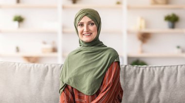 Mutlu, olgun, Müslüman bir kadın tesettüre oturmuş kameraya gülümsüyor. Modern Orta Doğu Arap Ev Kadınları Salonda Poz Veriyor