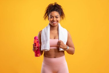 Antrenman kıyafeti giyen ve elinde pembe bir su şişesi tutan genç bir kadın kendinden emin duruyor. O, boynuna bir havlu sararak gülümsüyor, vücut bulmuş sağlık ve vücut bulmuş..