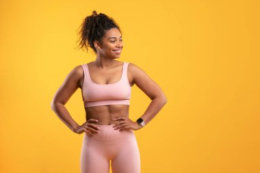 Genç bir kadın tebessüm ediyor, spor kıyafetini gösteriyor. Parlak sarı arka plan, sağlıklı bir yaşam tarzı ve kendine güveni vurgulayarak, onun tonlu fiziğinin ve pozitif enerjisinin altını çiziyor.