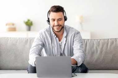 Uzaktan öğrenme konsepti. Kulaklık takan, dizüstü bilgisayarla çalışan, evdeki koltukta oturan, fotokopi çeken bir adam. Yakışıklı adam kanepede oturup Webinar izliyor, online eğitimin tadını çıkarıyor.
