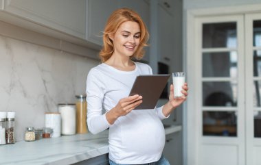 Hamile bir kadın parlak bir mutfakta elinde bir bardak süt tutarken elindeki tablete bakarken gülümsüyor. Konforlu ortamda modern dekor ve doğal ışık yer alıyor..