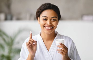 Beyaz bornoz giyen neşeli bir kadın bir elinde deri kavanozu, bir elinde de gülümseyerek parmağıyla işaret ediyor. Sıcak bir banyoda, sabah kendi kendine bakım ritüelinin tadını çıkarıyor..