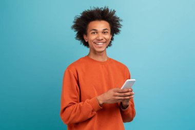 Kıvırcık saçlı genç bir Afrikalı Amerikalı canlı mavi bir fon karşısında duruyor. Akıllı telefonu tutarken neşeli bir şekilde gülümsüyor, rahat bir yaşam tarzı anı yaşıyor..