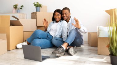 Sevgi dolu siyah çift dizüstü bilgisayarlarında aileleriyle görüntülü görüşme yapıyorlar ve kameraya el sallıyorlar, yeni evlerinde yerde oturuyorlar. Yeni evliler kendi evlerine taşınıyor.