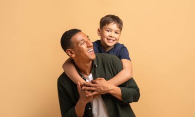 Bir baba, her ikisi de gülücüklerini paylaşarak oğlunu şakacı bir şekilde kucaklar. Güçlü bir bağ sergiliyorlar. Sıcak, izole bir ortamda mutluluk saçıyorlar. Aile sevgisini kutluyorlar..