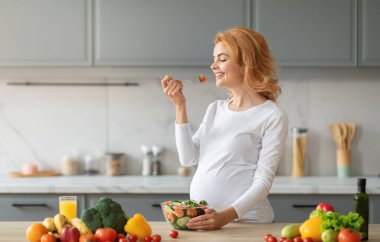 Gülümseyen hamile bir kadın, günümüzün mutfak ortamında renkli sebzeler ve meyvelerle çevriliyken çatal dolusu taze salatanın tadını çıkarıyor..