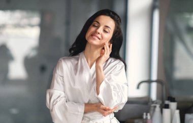 Esmer saçlı beyaz bir kadın şık banyosunda kendini şımartırken gülümsüyor. Cilt bakımı rutiniyle meşgul. Rahatlama ve kendine bakım hissi yayıyor..