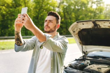 Bağlantı yok, araba arızası, kötü mobil ağ konsepti. Kırık otomobilin yanında duran, cep telefonuyla el kaldıran, yol kenarından yardım çağırmaya çalışan zavallı bin yıllık adam başarısız oldu.