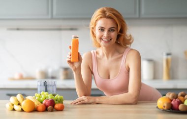 Neşeli bir kadın mutfağında durmuş taze sıkılmış meyve suyu şişesini gösteriyor. Tezgahın etrafında sağlıklı bir yaşam tarzını ön plana çıkaran çeşitli meyveler var..