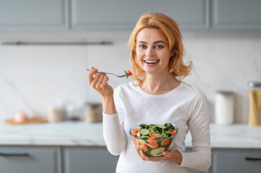 Sarı saçlı hamile bir kadın şık bir mutfakta gülümserken bir kase salata tutuyor. Bir ısırık almaya hazırlanırken mutlu görünüyor, sağlıklı bir yaşam tarzı ve taze malzemeler sergiliyor..
