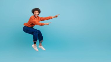 Kıvırcık saçlı genç bir Afrikalı Amerikalı parlak mavi bir arka plana karşı heyecanla zıplıyor. Turuncu bir kazak ve kot pantolon giyiyor, canlı bir atmosfer yaratıyor, boşluğu kopyalıyor.