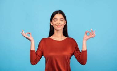 Bin yıllık esmer bir kadın sakin bir ifadeyle meditasyon yapar. Mavi bir arkaplanda, sağlığını ve huzurunu arttırmak için farkındalık uyguluyor..