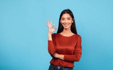 Uzun esmer saçlı bir kadın gülümsüyor ve iyi bir el hareketi yapıyor. O sade, şık bir elbise giyer ve canlı mavi bir arkaplan karşısında güvenle durur..