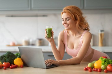 Yeşil smoothie içerken kızıl saçlı genç bir kadın gülümsüyor. Etrafı renkli sebzeler ve taze ürünlerle çevriliyken mutfak tezgahında dizüstü bilgisayar kullanıyor..