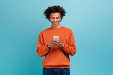 Kıvırcık saçlı genç bir Afrikalı Amerikalı akıllı telefonuyla parlak mavi bir arka plana mesaj atarken gülümsüyor. Turuncu bir kazak ve kot pantolon giyiyor..