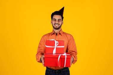 Parti şapkalı bir Orta Doğulu elinde parlak sarı bir arka planla kırmızı hediye kutularını tutarken gülümsüyor. Neşeli ifadesi kutlama ve başarıyı yansıtıyor..