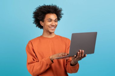 Bin yıllık kıvırcık saçlı bir adam dizüstü bilgisayarıyla meşgul, özgüvenle gülümsüyor. Günlük turuncu bir kazak giyiyor, pozitif ve çekici bir atmosfer yaratıyor..