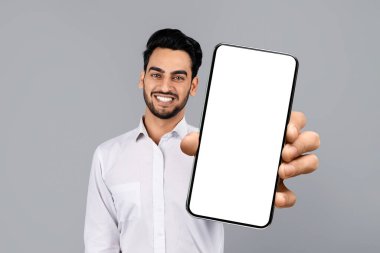 Orta Doğulu bir işadamı stüdyoda güvenle duruyor ve dostça bir gülümsemeyle boş bir akıllı telefon ekranı sergiliyor. Bu an, modern girişimciliğin özünü yakalar..