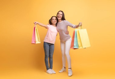 Bir anne ve genç kızı, parlak sarı bir arka planda renkli alışveriş torbaları taşımaktan mutluluk duyarlar. Birbirlerine uyumlu gülümsemeleri, gezileri sırasında ortak bir neşe ve şefkat anını yansıtıyor..