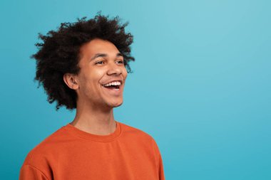 Kıvırcık saçlı neşeli, Afro-Amerikan bir adam neşeyle gülüyor. Günlük turuncu bir süveter giyiyor ve parlak mavi bir arka planın önünde duruyor.