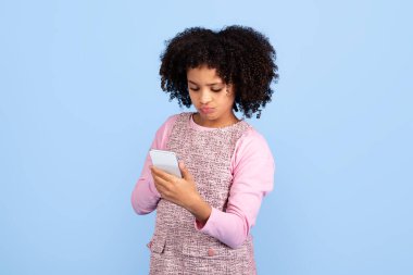 Genç, neşeli bir zenci kız mavi bir arkaplanın önünde telefonuyla meşgul bir şekilde dikiliyor. Kıvırcık saçları yüzünü çerçeveler, mutluluğunu ve enerjik enerjisini yansıtır..