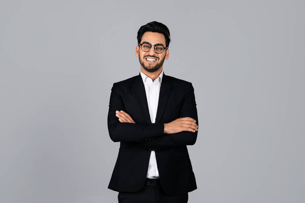 A young Arab entrepreneur stands with arms crossed, wearing a formal black suit and glasses. The neutral backdrop highlights his confident smile and professional demeanor in a studio environment.