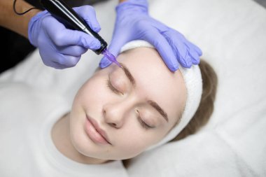 Bir kadın kalıcı bir makyaj ameliyatı olurken güzellik salonunda dinlenir. Güzellik uzmanı, bu uzun süreli seans süresince müşterinin doğal güzelliğini artırmak için ustalıkla makyaj yapar..